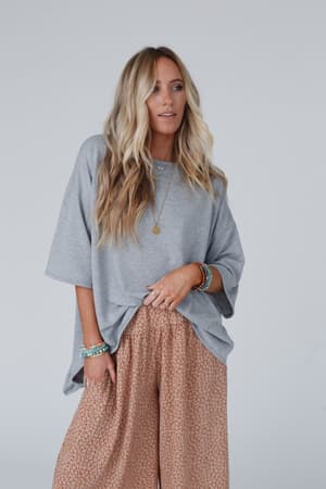 A woman with long, wavy blonde hair wears The Nest Must Have High Low Tee in Heather Gray and wide-legged peach patterned pants, accessorized with boho bracelets and a necklace, standing against a plain light backdrop.