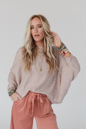 A woman with long blonde hair wears The Nest Fly With Me Batwing Sleeve Knit Sweater in Sand and pink drawstring pants, accessorized with layered necklaces and colorful beaded bracelets, posing before a plain light background.