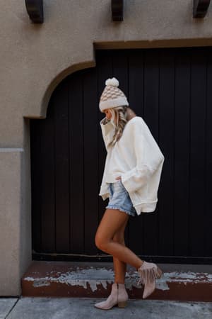 A person wearing the Dana Oversized Diamond Texture Sweater in Ivory, a beige knit hat, denim shorts, and tan fringe ankle boots stands on a sidewalk in front of a dark wooden door, looking down with one hand near their face.