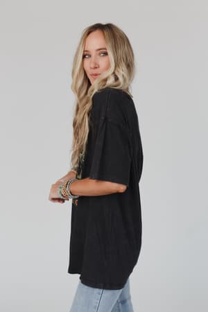 A woman with long, wavy blonde hair stands sideways against a plain light background, smiling slightly while wearing the Share The Love Graphic Tee in charcoal and light blue jeans. She has bracelets on her left wrist.