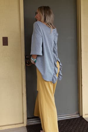A woman smiles at a door, wearing The Nest Must Have High Low Tee in Heather Gray with ruffle details down the back and wide-leg yellow pants. She accessorizes with bracelets, and her blonde hair is worn down.