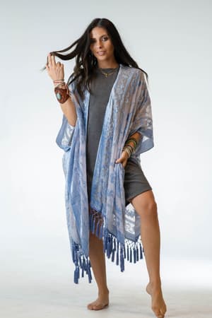 A woman with long dark hair stands barefoot on a white background, wearing a short gray dress and the Crushin' On You Velvet Kimono in light blue, featuring tasseled edges. She accessorizes with layered jewelry and holds part of her hair.