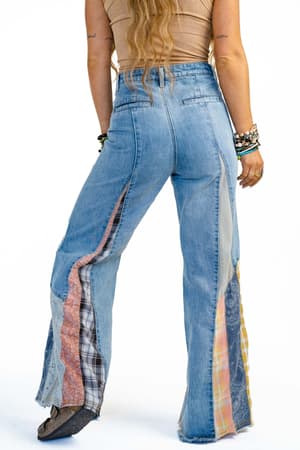 A person with long hair, wearing a tan top and Magnolia Patchwork Jeans - Washed Denim with blue flared legs and multicolored patchwork panels, stands facing away. They wear bracelets on both wrists and have visible arm tattoos.