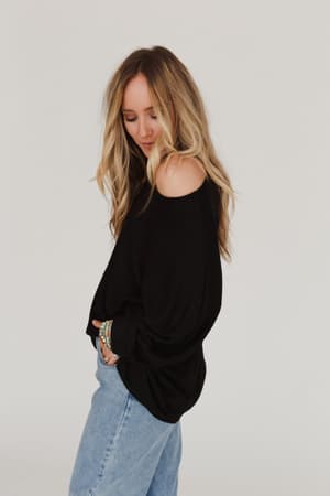 A woman with long blonde hair stands in profile against a plain white background, smiling gently and wearing the Fly With Me Batwing Sleeve Knit Sweater in black with light blue jeans.