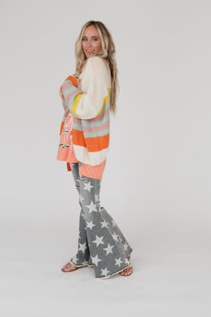 A woman with long blonde hair stands sideways, smiling. She wears the Retro Babe Striped Cardigan - Cream over a coral shirt, gray bell-bottom pants with white stars, and sandals, set against a plain white background.