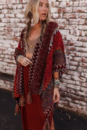 A woman with long blonde hair styled in loose braids wears the Desert Dreams Crochet Ruana - Maroon from her boho closet, along with layered necklaces, bracelets, and a burgundy skirt, standing by a white brick wall.