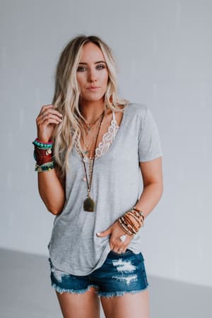 A woman with long blonde hair wears the Scoop Neck Bralette Tee - Gray and ripped denim shorts, accessorized with layered necklaces, colorful bracelets, and rings, standing against a plain light background.