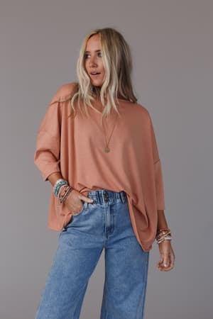 A woman with long blonde hair wears The Nest Cozy Cool Oversized Tee in Coral, blue high-waisted jeans, layered bracelets, and a pendant necklace, standing against a plain gray background.