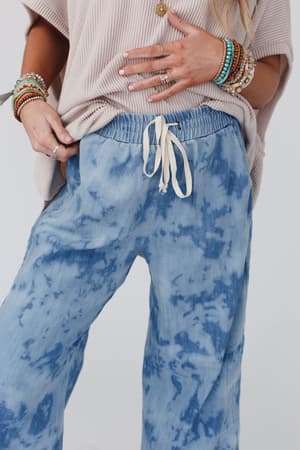 A person wears The Nest So Comfy Wide Leg Denim Pant - Bleach Dye Denim with a loose beige top and colorful beaded bracelets and rings, showing only their lower torso and hands.