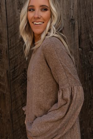 A woman with long blonde hair wears The Nest Sweet Heart Balloon Sleeve Knit Sweater in mocha, standing before a wooden wall and smiling as she looks to the side.