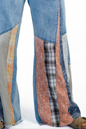 A person wears Magnolia Patchwork Jeans - Washed Denim, featuring colorful plaid, paisley, and checkered panels with a frayed hem, paired with brown shoes. Only the lower half of the legs is visible against a white background.