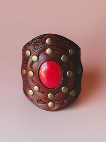 A wide leather cuff bracelet shot close-up against a dusty rose background — rich brown tooled leather with brass studs circling a bold red stone center, the kind of piece that looks like it has a story.