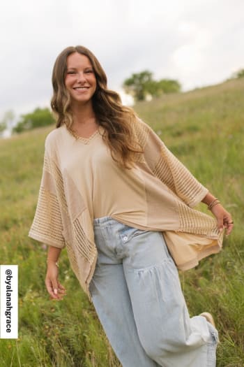 A woman with long brown hair smiles in a grassy field, looking relaxed and happy in her boho style. She wears the City Drifter Oversized Contrast Top - Taupe paired with light blue wide-leg jeans under a partly cloudy sky.