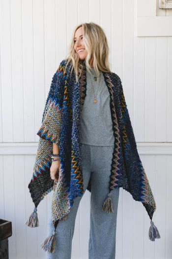A woman with long blonde hair models the Desert Dreams Crochet Ruana - Cobalt, featuring blue and multicolored knit fabric with tassels, worn over a light gray outfit—a perfect boho piece—while smiling in front of a white paneled wall.