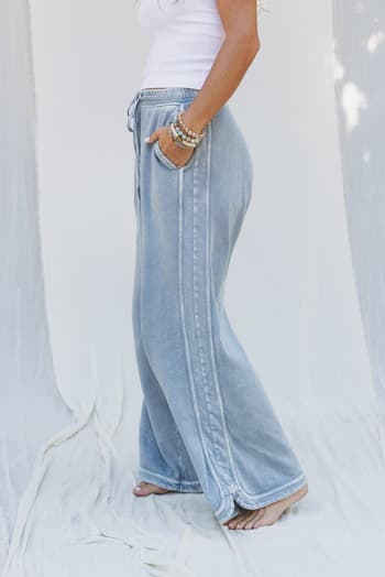 A woman stands barefoot against a white fabric backdrop, wearing the So Comfy Wide Leg Pant Cropped Length in Faded Blue with a high-rise elastic waist and a white tank top, one hand in her pocket and bracelets on her wrist.