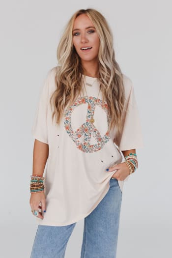 A woman with long wavy blonde hair wears the Jude Oversized Peace Sign Graphic Tee in oatmeal, paired with blue jeans and colorful beaded bracelets, standing against a plain light background.