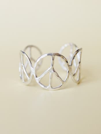 A wide silver cuff bracelet with repeating peace sign cutouts arranged in an open, sculptural band — shot close up against a warm cream background with soft, even light.