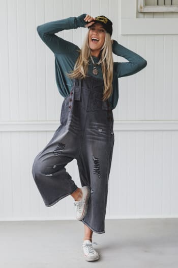 A smiling woman playfully poses on one foot in a black cap, teal long-sleeve shirt, and High Hopes Slouchy Denim Overalls - Black, featuring distressed details. She completes the look with white sneakers against a white paneled wall.