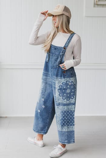 A woman wearing the High Hopes Slouchy Denim Overalls - Denim Bandana, a long-sleeve white shirt, white sneakers, and a beige baseball cap stands indoors smiling as she touches her hat.
