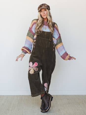 A full-body model shot of a layered boho outfit built around olive corduroy overalls with patchwork flower appliqués, worn over a multicolor stripe knit sweater and finished with a plaid newsboy cap and leopard print boots — the kind of look that fee
