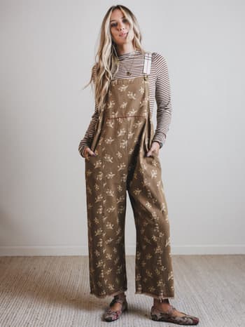 Woman wearing a patterned brown jumpsuit with a striped shirt underneath, standing against a plain white wall.