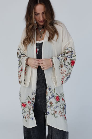Wearing the Fun In Florals Kimono - Cream, a woman with long wavy hair and layered necklaces stands looking down, adjusting its sheer floral-embroidered fabric over her dark outfit.