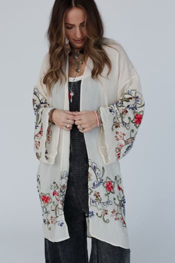 Wearing the Fun In Florals Kimono - Cream, a woman with long wavy hair and layered necklaces stands looking down, adjusting its sheer floral-embroidered fabric over her dark outfit.