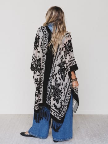 A woman with long, wavy hair faces away, wearing the Crushin' On You Velvet Kimono - Black over wide-leg denim pants and black shoes, standing in a minimalist room with a plain white wall.