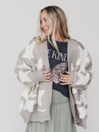 A smiling woman with long blonde hair wears the Wrapped In Stars Oversized Cardigan - Taupe, featuring white star patterns, over a black graphic T-shirt and light green pleated skirt. She completes her look with several rings.