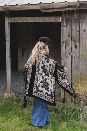 A person with long blonde hair, a black hat, the *Crushin' On You Velvet Kimono - Black*, and wide-leg jeans stands near an old wooden building with weathered walls and a metal roof.