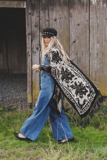 A woman in wide-leg jeans, black flats, a black cap, and the Crushin' On You Velvet Kimono - Black walks on grass before a weathered wooden wall.