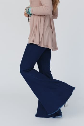A woman wearing a long-sleeved beige top and The Signature Flare - Dark Denim stands against a plain light background, accessorized with layered bracelets. Her face is not visible in the image.