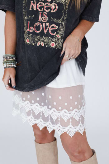 A person wears the Layering Love Lace Slip - Ivory under a white polka dot skirt, paired with beige knee-high boots and multiple bracelets. Only their lower half is visible.