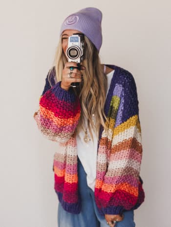 A woman with long blonde hair wears a purple beanie and the Wrapped up in Color Knit Cardigan - Rainbow as she smiles, holding a vintage film camera to her face against a plain white background.