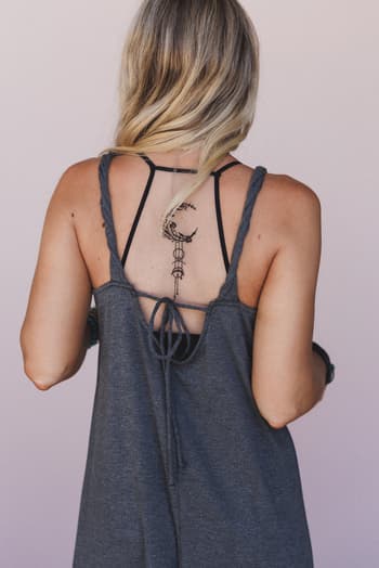 A woman with blonde hair models the Crescent Moon Tattoo Bralette – Black, featuring a black lace design and a geometric crescent moon tattoo print on her upper back, visible under a dark gray sleeveless open-back top.