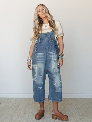 A model in slouchy cropped denim overalls with bandana-print patchwork and distressing, styled over a vintage-striped tee with brown platform clogs and stacked beaded bracelets — effortless and a little worn-in, like your coolest thrifted find.