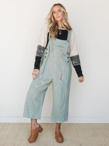 Wide-leg distressed denim overalls in a faded sage wash, layered over a mixed-print long sleeve top and finished with tan studded clogs — effortlessly lived-in and the kind of outfit that looks like you've had it forever.