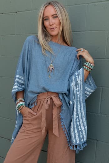 A woman with long blonde hair poses against a green brick wall, wearing the Fallon Embroidered Sleeve Tassel Poncho in blue, featuring geometric white embroidery and tassels, paired with tan high-waisted pants and layered accessories.