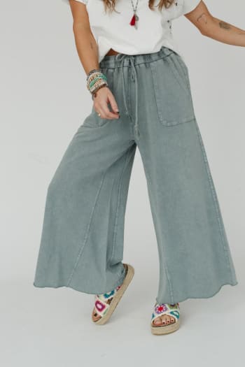 Wearing a white t-shirt, Relaxing Robin Wide Leg Pant in Faded Teal, and colorful embroidered sandals, this person also accessorizes with bracelets and a necklace. Only their lower face and body are visible.