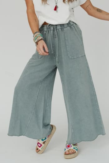 Wearing a white t-shirt, Relaxing Robin Wide Leg Pant in Faded Teal, and colorful embroidered sandals, this person also accessorizes with bracelets and a necklace. Only their lower face and body are visible.