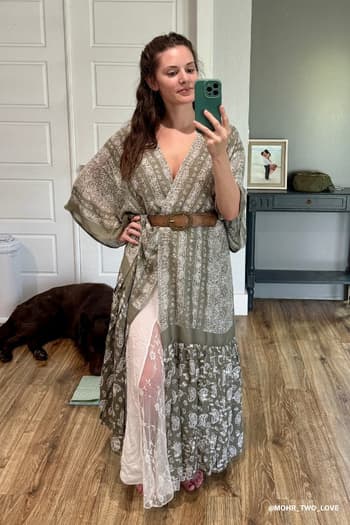 A woman wears the Paisley Tapestry Free Flow Duster Kimono - Olive and takes a mirror selfie on wooden floors, with a brown dog lying on a mat behind her and a desk with framed photo and decor in the background.