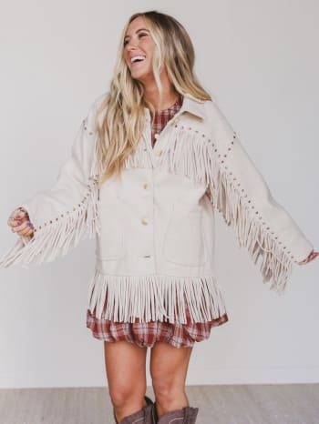 Woman wearing a beige fringed jacket over a plaid dress on a plain background