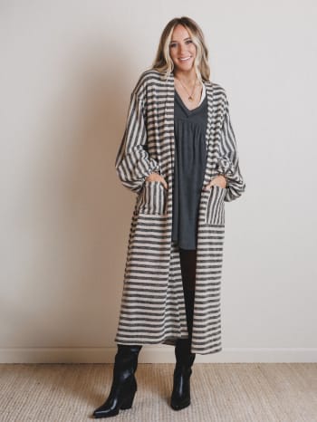 Woman wearing a long black and white striped coat over a dark dress, standing against a plain wall.