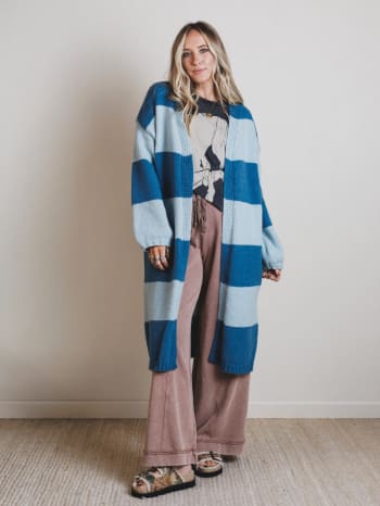 Woman wearing a long blue and gray striped cardigan with brown pants against a plain background