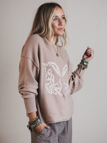Woman wearing a beige sweater with a horse design against a plain background