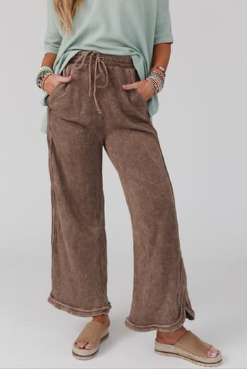 A person models the So Comfy Wide Leg Pant Full Length in Mocha, standing with hands in pockets. Only their lower torso, legs, and arms are visible against a plain background.