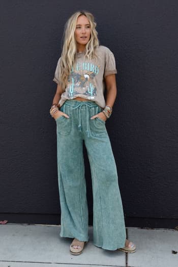 A woman poses against a dark wall in the Relaxing Robin Wide Leg Pant in New Teal, paired with a graphic tee and sandals. Her long blonde hair and stacked bracelets complete the laid-back look, with her hands resting in her pockets.