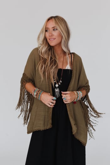 A woman with long blonde hair wears a black dress and the Dreamy Macramé Kimono in Olive, styled with colorful beaded bracelets and necklaces, standing against a plain light background.