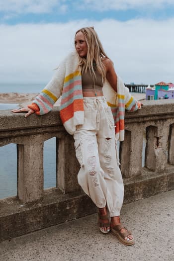 A woman leans against a concrete railing by the water, wearing a beige tank top, white ripped cargo pants, sandals, and the Retro Babe Striped Cardigan - Cream. Colorful buildings and a beach can be seen in the background.