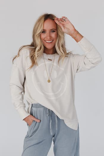 A woman with long blonde hair smiles at the camera, wearing the Catch My Smile Pullover Top in khaki and loose blue pants. One hand is in her pocket while the other is raised near her head.